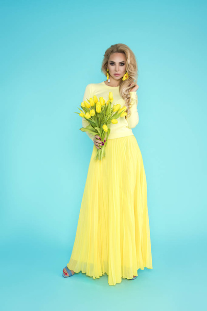 Spring Woman. Beauty Summer model girl with colorful clothes, holding a bouquet of spring flowers. Beautiful lady with yellow tulips on the blue background. - Fotó, kép