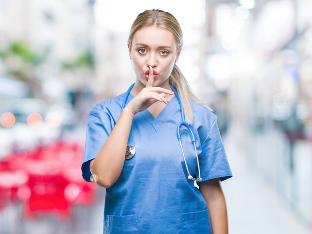 Νεαρή ξανθιά χειρουργός γιατρός γυναίκα πέρα από το απομονωμένο υπόβαθρο ζητώντας να είναι ήσυχη με το δάχτυλο στα χείλη. Σιωπή και μυστικό έννοια. - Φωτογραφία, εικόνα