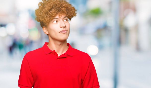 Nuori komea mies afro hiukset yllään punainen t-paita hymyillen näköinen puolella ja tuijottaen pois ajattelu
. - Valokuva, kuva