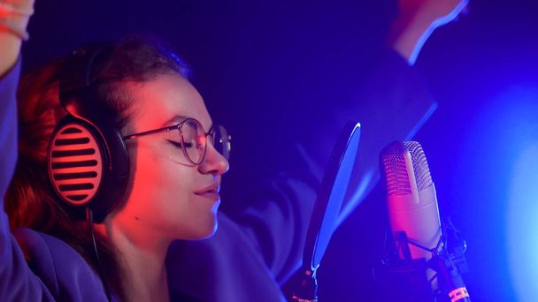 Молодая женщина в очках поет в неоновом свете в студии с поднятыми руками
 - Фото, изображение