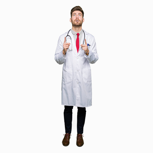 Молодий красивий лікар чоловік в медичному пальто здивований і здивований, дивлячись вгору і вказуючи пальцями і підняті руки
. - Фото, зображення