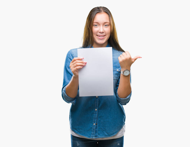 Mulher branca jovem segurando folha de papel em branco sobre fundo isolado apontando e mostrando com o polegar até o lado com o rosto feliz sorrindo
 - Foto, Imagem