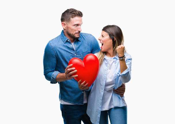 Young couple in love holding red heart over isolated background annoyed and frustrated shouting with anger, crazy and yelling with raised hand, anger concept - Fotografie, Obrázek