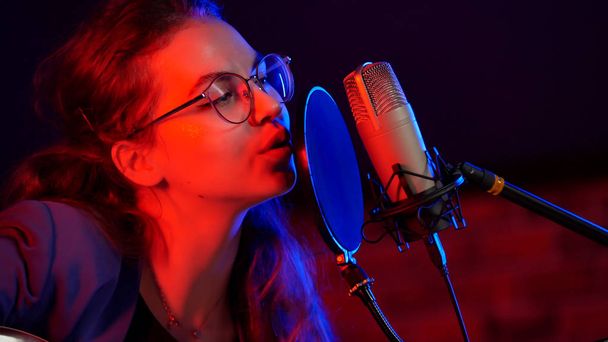 Egy fiatal nő, kerek szemüveg, neon világítás a sötét stúdióban énekel - Fotó, kép