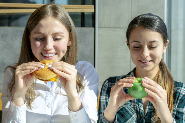 dvě krásné dívky v kavárně jíst hamburger a apple, zdravé a nezdravé jídlo - Fotografie, Obrázek