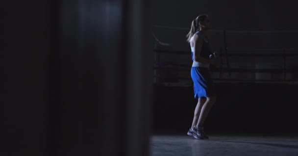 Кардиотренировка для бойцов. Молодая красивая женщина профессиональный боксер в тренировочном зале прыгает на веревке в свете контрас в темной комнате в синей одежде
 - Кадры, видео