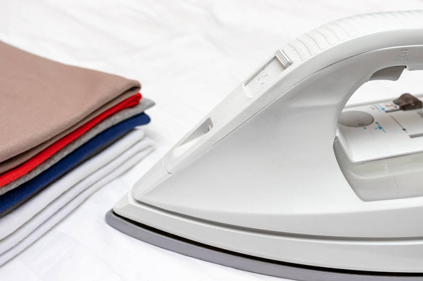 Modern electric iron and a pile of clothes on white background close up - Photo, Image