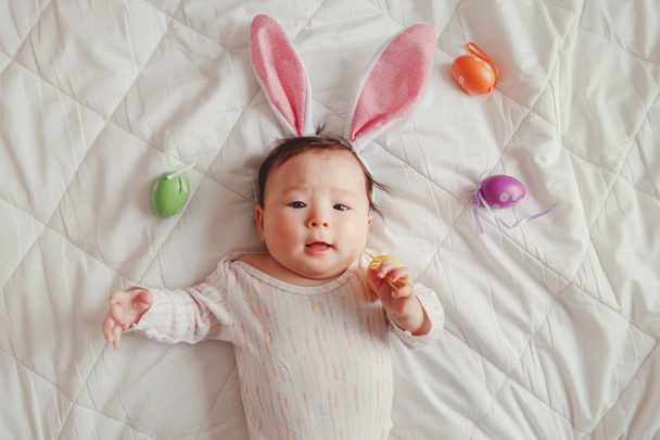 Lindo adorable bebé asiático mixto con orejas de conejo de Pascua rosa acostado en la cama en el dormitorio con huevos de colores. Niño divertido infantil celebrando la fiesta cristiana tradicional. Vista desde arriba
 - Foto, imagen