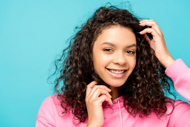 smiling african american young woman touching curly hair isolated on blue  - Photo, Image