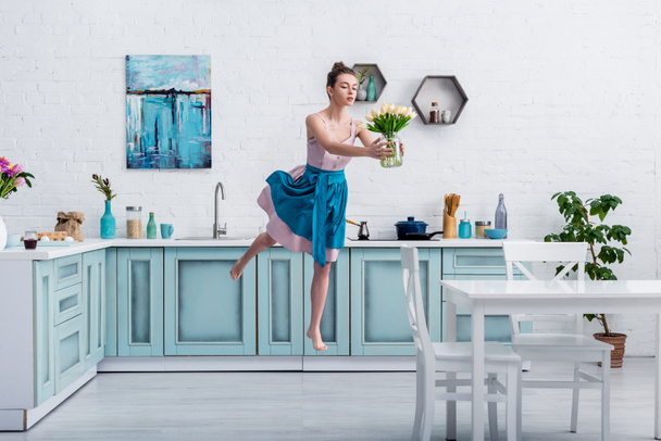 beautiful barefoot girl in elegant dress and apron flying in air with bouquet of tulips in glass jar in kitchen - Photo, Image