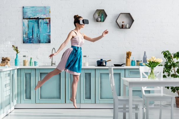 junge barfüßige Frau fliegt in Virtual-Reality-Headset durch die Luft und gestikuliert in der Küche - Foto, Bild