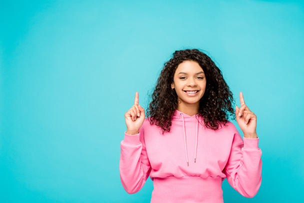 happy curly african american girl pointing with fingers and looking at camera isolated on blue  - Foto, afbeelding