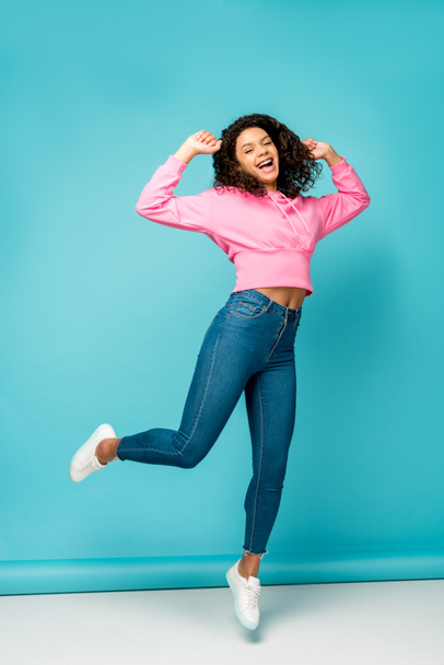 happy curly african american girl in jeans jumping on blue - 写真・画像