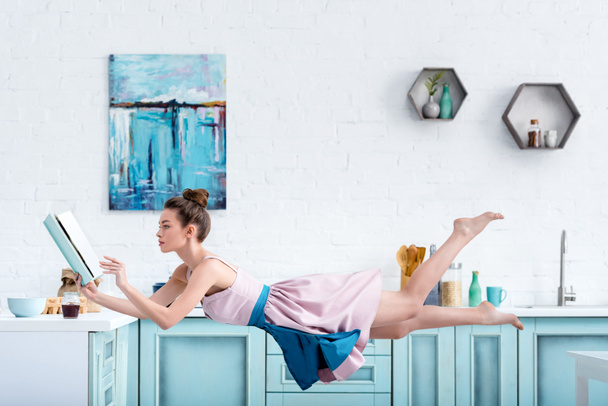 junge konzentrierte barfüßige Frau schwebt in der Luft, während sie Rezept in Kochbuch liest - Foto, Bild