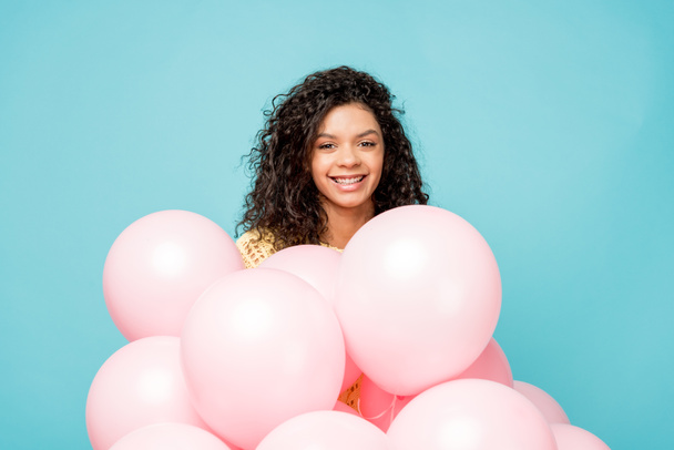happy curly african american girl near pink air balloons isolated on blue - Foto, afbeelding