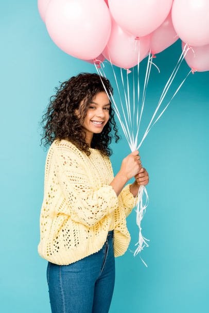 attractive curly african american girl holding pink air balloons on blue  - Zdjęcie, obraz
