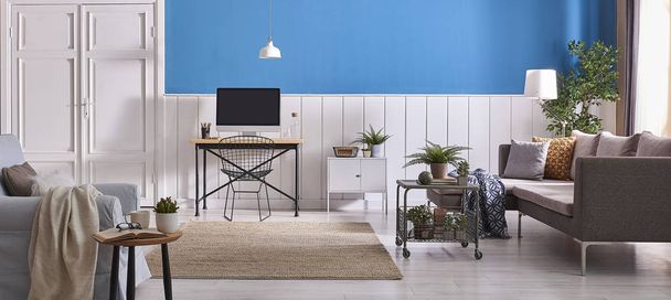 Sala de estar moderna, fundo de parede branco e azul, mesa de trabalho de madeira, sofá cinza, computador e poltrona azul. Lâmpada branca e plantas verdes
 - Foto, Imagem
