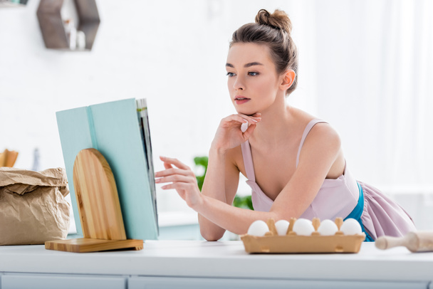 attractive girl reading recipe in cookbook with bakery ingredients around  - Foto, Imagem