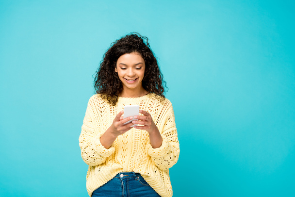 όμορφη σγουρά αφρικανική Αμερικανίδα γυναίκα χαμογελά ενώ χρησιμοποιεί smartphone απομονώνεται σε μπλε  - Φωτογραφία, εικόνα