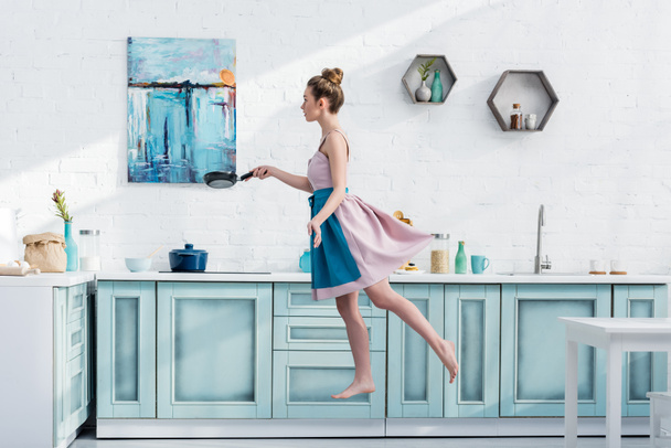 attractive barefoot young woman in apron levitating in kitchen while holding pan  - Valokuva, kuva