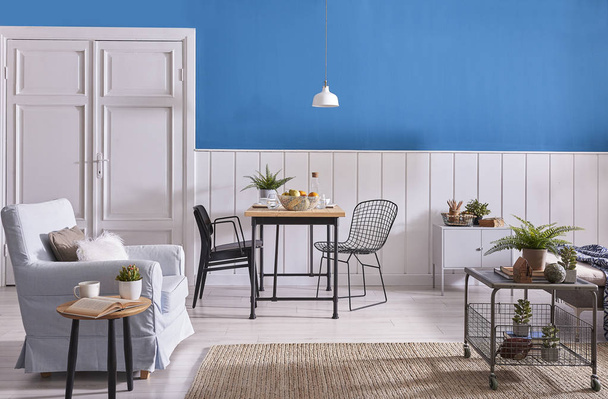 Modern yemek odası dekorasyon, ahşap masa ve siyah sandalye. Mavi ve beyaz duvar arka plan. Klasik duvar detay. Gri mobilya, orta masa, kanepe ve mavi koltuk. Hasır halı ve beyaz lamba. - Fotoğraf, Görsel