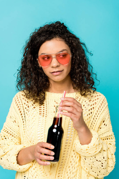 attraente ragazza afro-americana in occhiali da sole bottiglia di tenuta con paglia isolata su blu
 - Foto, immagini
