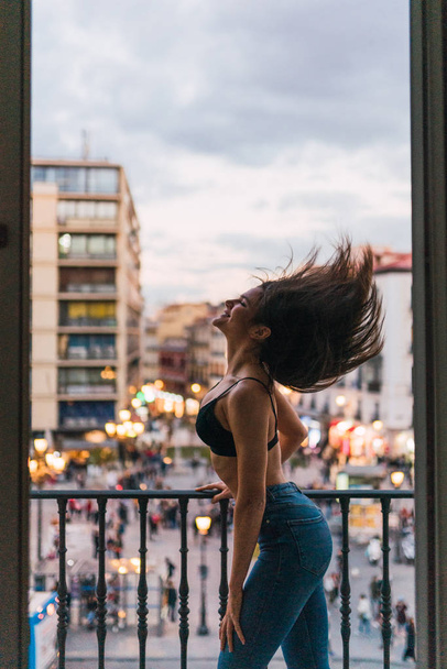 Zijaanzicht van slanke brunette dame met winderige haren draagkracht op balkon met uitzicht op de stad stree - Foto, afbeelding
