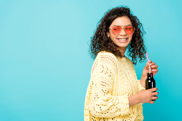 happy curly african american girl in sunglasses holding bottle with straw isolated on blue - Photo, Image