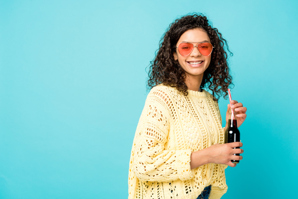 uśmiechnięta kręcone African American Girl w okulary trzymając butelkę ze słomką na niebiesko - Zdjęcie, obraz
