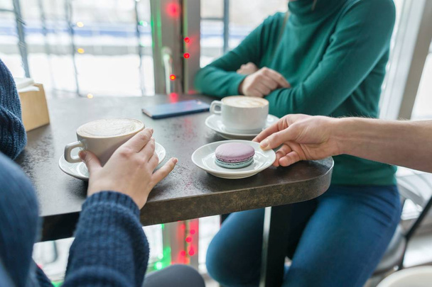 Primer plano de dos manos de las mujeres con tazas de café y macarrones en el plato en la mano del hombre en la mesa de piedra gris, fondo de la cafetería
 - Foto, Imagen