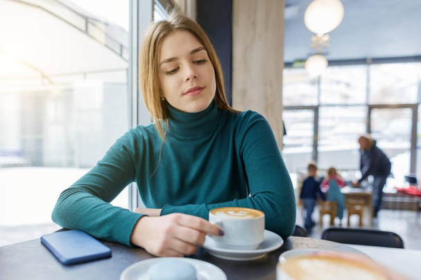 Young beautiful girl with cup of coffee macaroons on plate, winter season in coffee shop - Foto, immagini