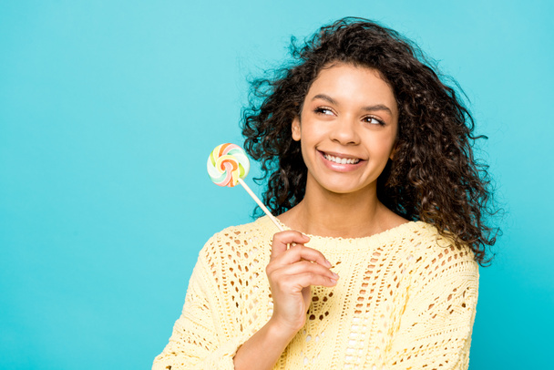 Happy Curly Afro-Amerikaanse meisje glimlachen terwijl lolly geïsoleerd op blauw - Foto, afbeelding