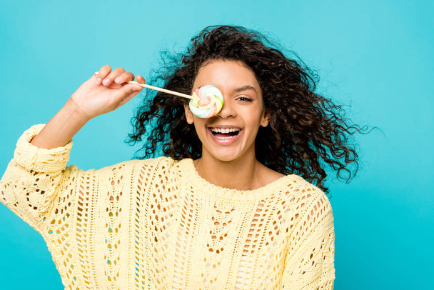 happy curly african american girl smiling while covering eye with lollipop isolated on blue - Φωτογραφία, εικόνα