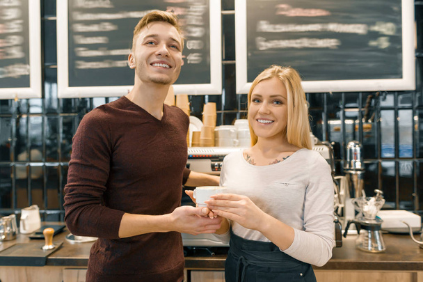 Happy smiling couple at the coffee shop counter, workers or owners with a cup of fresh art coffee looking at the camera. Small business, service, cafe - Photo, image