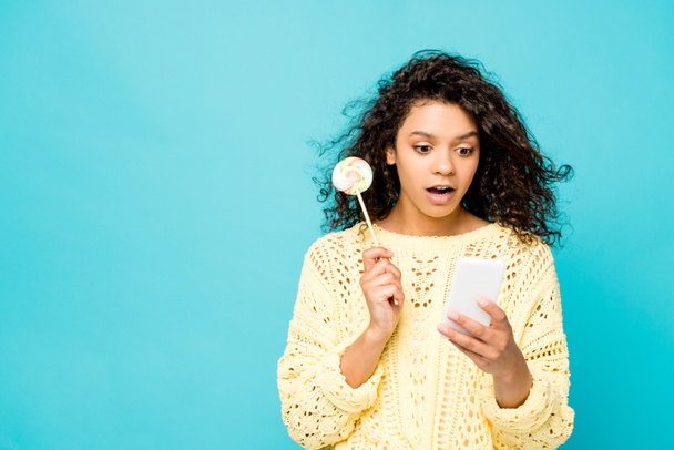 meglepett, göndör afro-amerikai lány gazdaságban nyalóka használata közben okostelefon elszigetelt kék - Fotó, kép