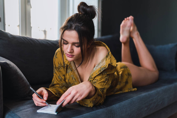 Young woman with pen and notebook lying on sofa in room - Foto, Imagen