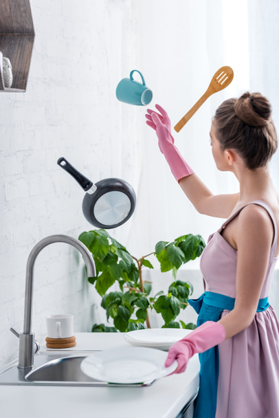 giovane donna in guanti di gomma guardando volare in aria utensili da cucina in cucina
 - Foto, immagini