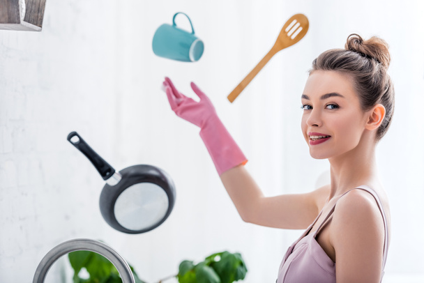 selective focus of young smiling woman in rubber glove pointing at flying in air cooking utensils in kitchen - Photo, Image