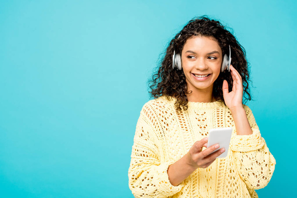 cheerful curly african american girl listening music in headphones while holding smartphone isolated on blue - Fotó, kép