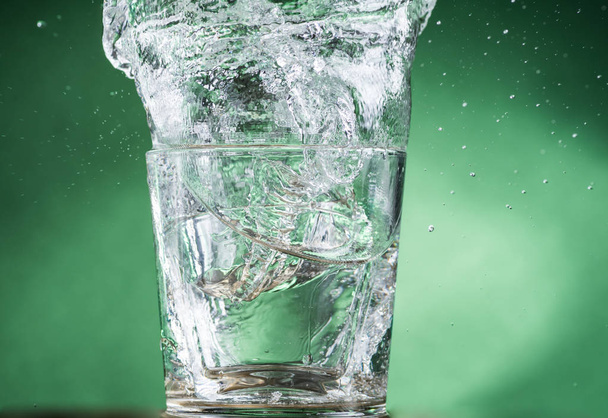 caindo pequeno vidro transparente com água clara e derramamento de água em um fundo verde
 - Foto, Imagem