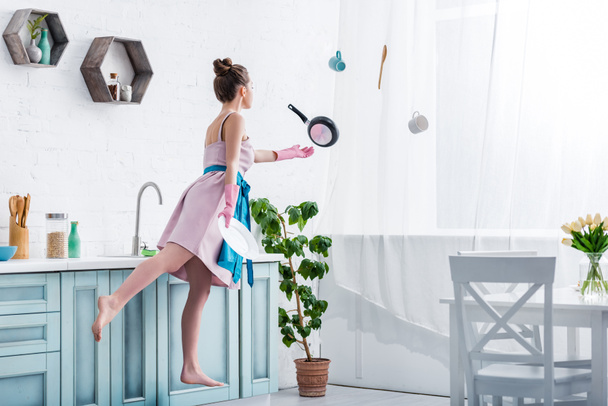 young woman in rubber gloves levitating in air with cooking utensils in kitchen  - Photo, Image