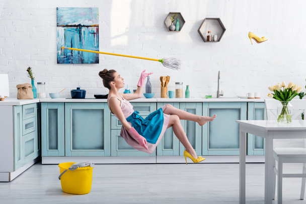Elegantes Mädchen in Schürze schwebt mit Wischmopp und gelbem Schuh beim Hausputzen in der Küche - Foto, Bild