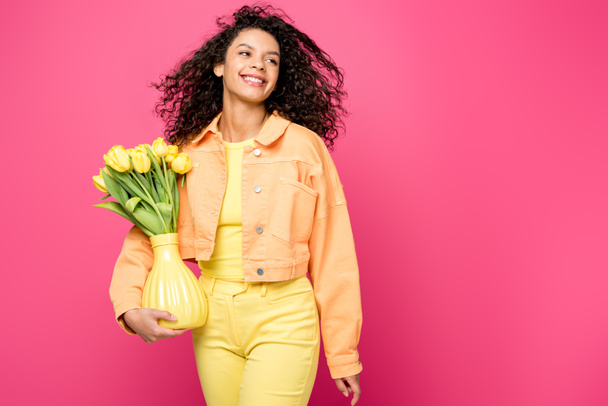happy african american girl holding vase with yellow tulips while standing isolated on crimson - Фото, изображение