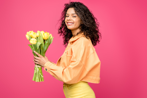 happy african american woman holding yellow tulips isolated on crimson - Zdjęcie, obraz