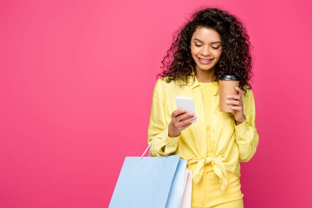 クリムゾンに分離された紙コップを保持しながら、スマートフォンを見てショッピングバッグを持つ幸せなアフリカ系アメリカ人の女性 - 写真・画像