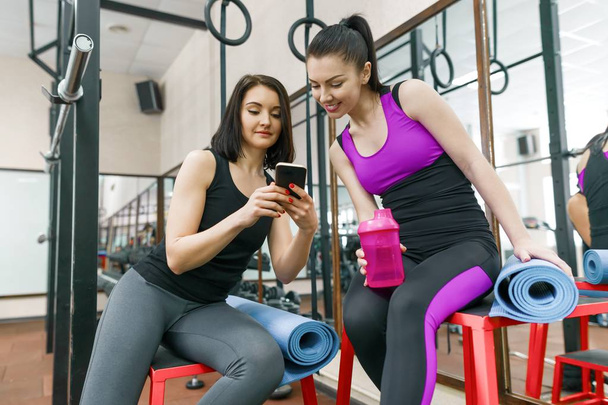 Dos jóvenes deportistas en el gimnasio hablando sonriendo con alfombras de fitness y mirando el teléfono móvil. Formación, trabajo en equipo, concepto de estilo de vida saludable
. - Foto, Imagen