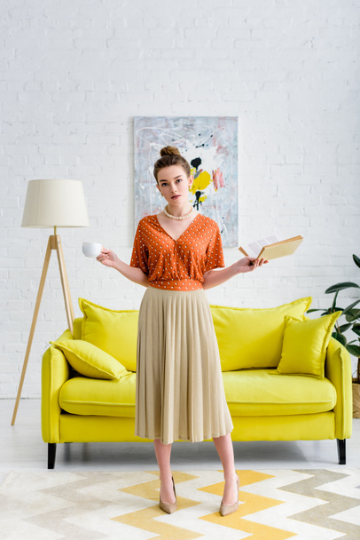 attractive elegant young woman holding book and coffee cup in living room - Photo, image