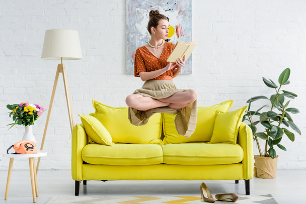 elegant young woman in lotus pose levitating in air while reading book in living room - Photo, Image