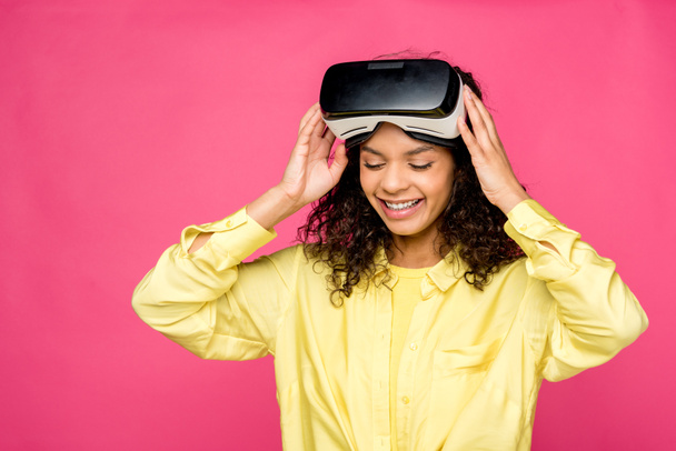 göndör afro-amerikai nő visel virtuális valóság fülhallgató és mosolyogva elszigetelt a bíbor  - Fotó, kép