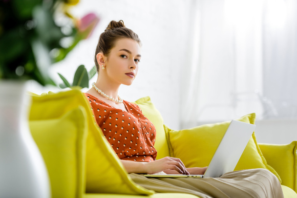 selective focus of fashionable young woman in pearl necklace sitting on yellow sofa and using laptop - Foto, Bild